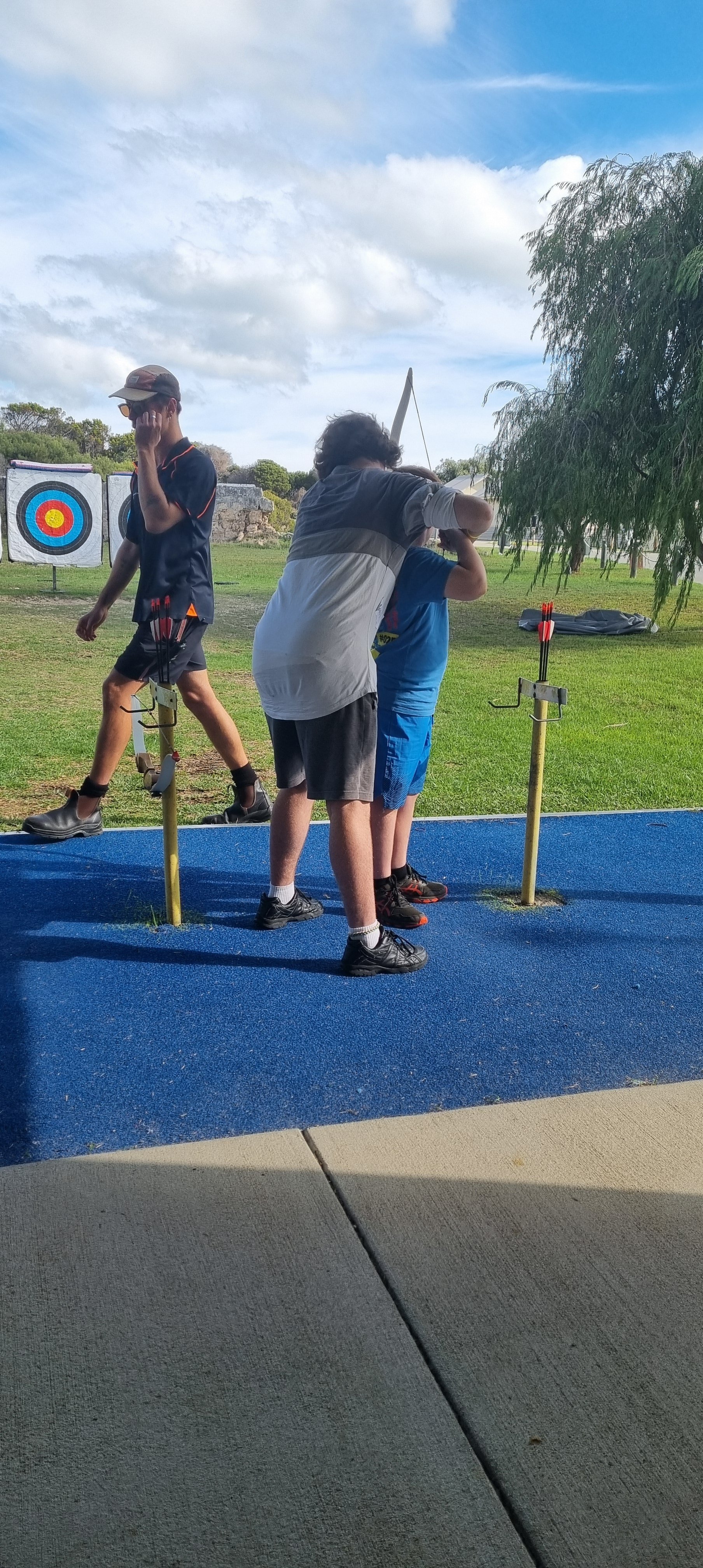 A group of children at an archery range, with one child receiving assistance from the other one while aiming a bow