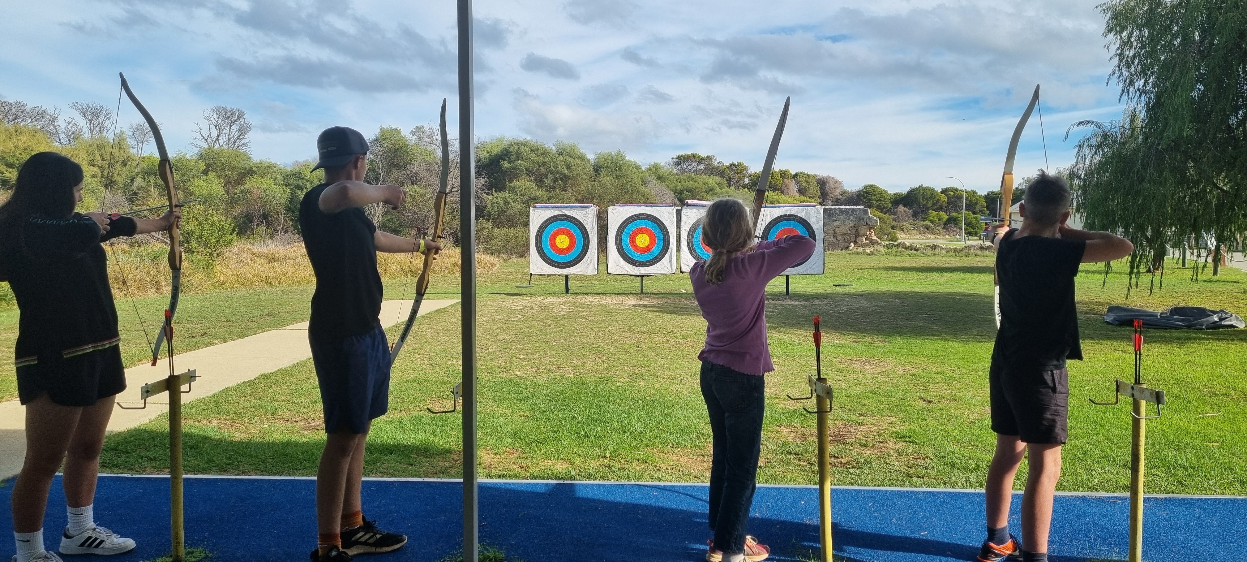 A group of kids at the archery range, each drawing back their bows while facing the targets on a grassy field