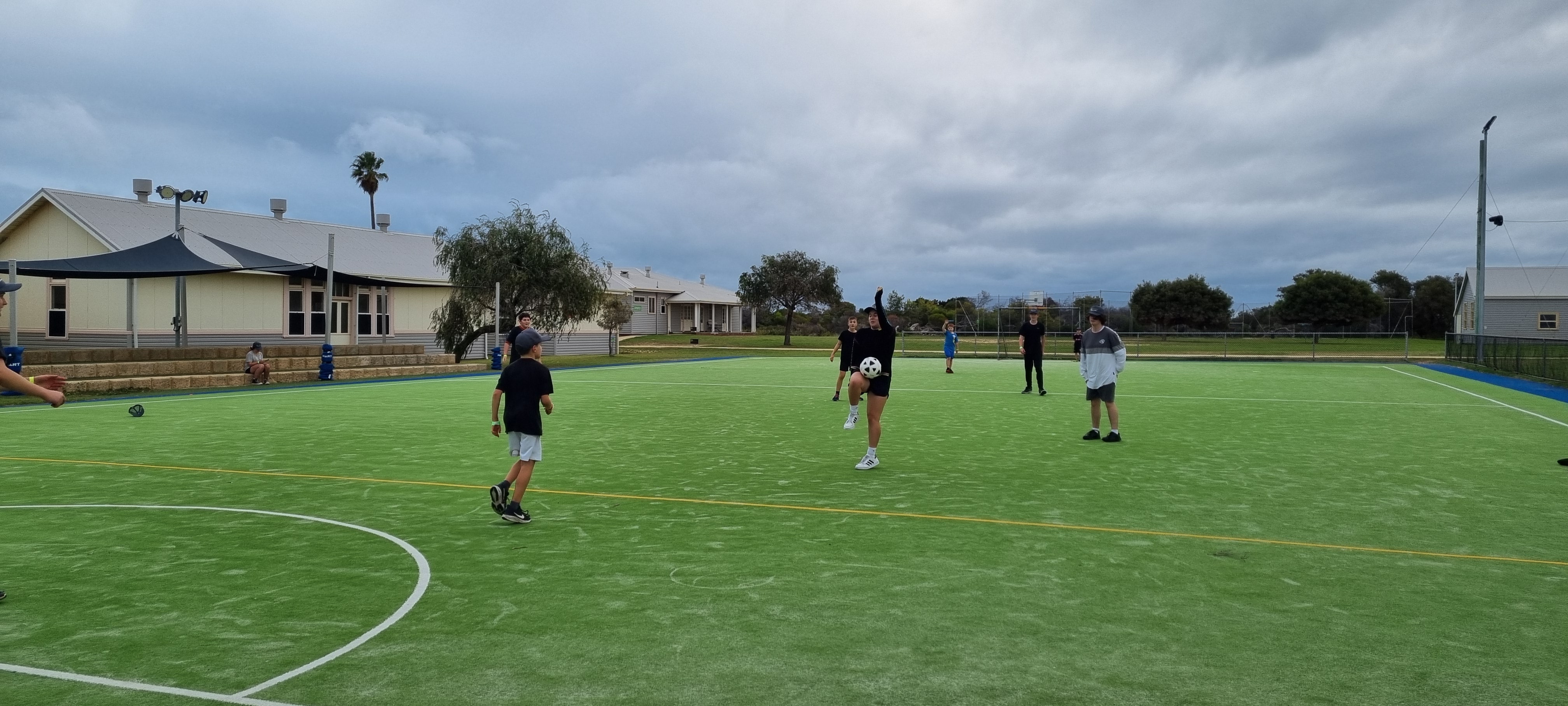 A group of children playing football on a grass field with a building and trees in the background