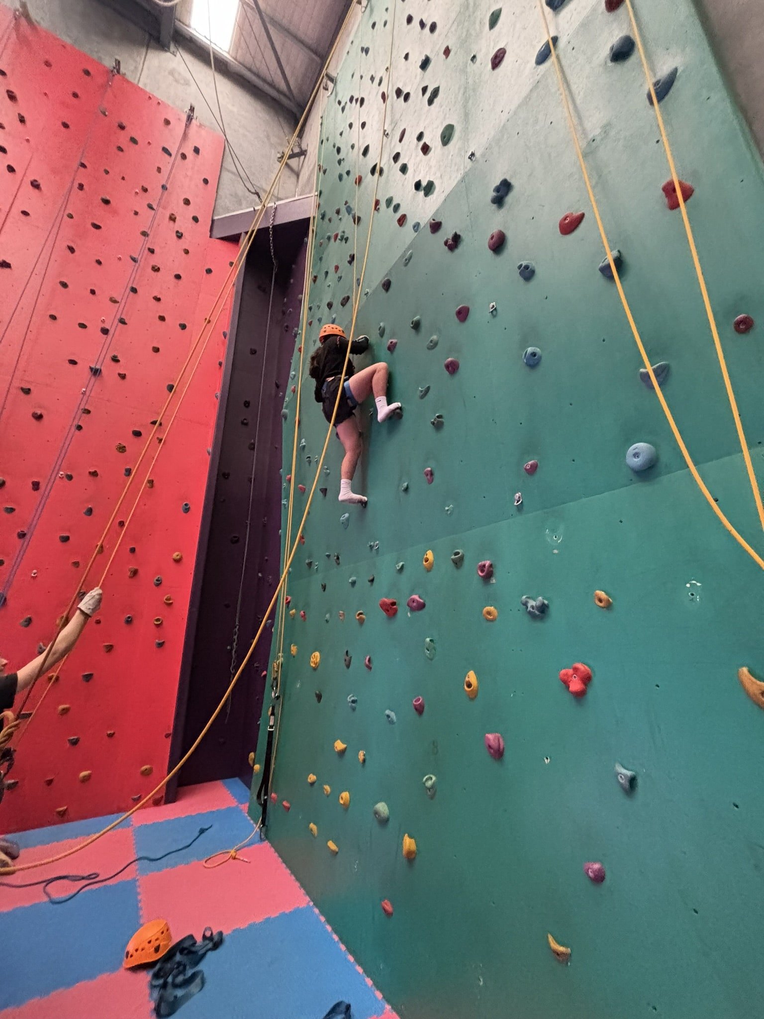 A child wearing a helmet and harness climbing a green section of the indoor rock wall