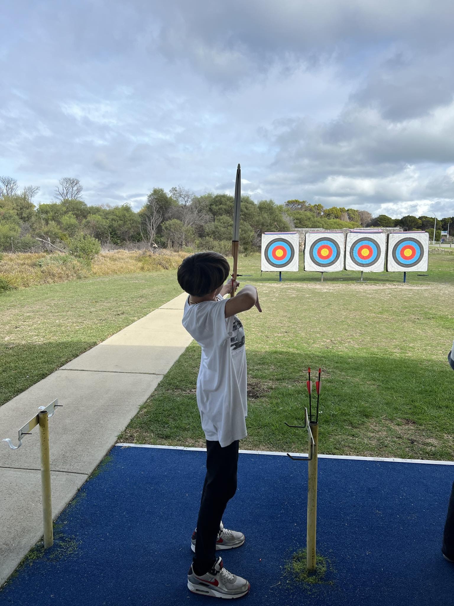 A child in a white t-shirt and black pants holding a bow, aiming at an archery target in the distance