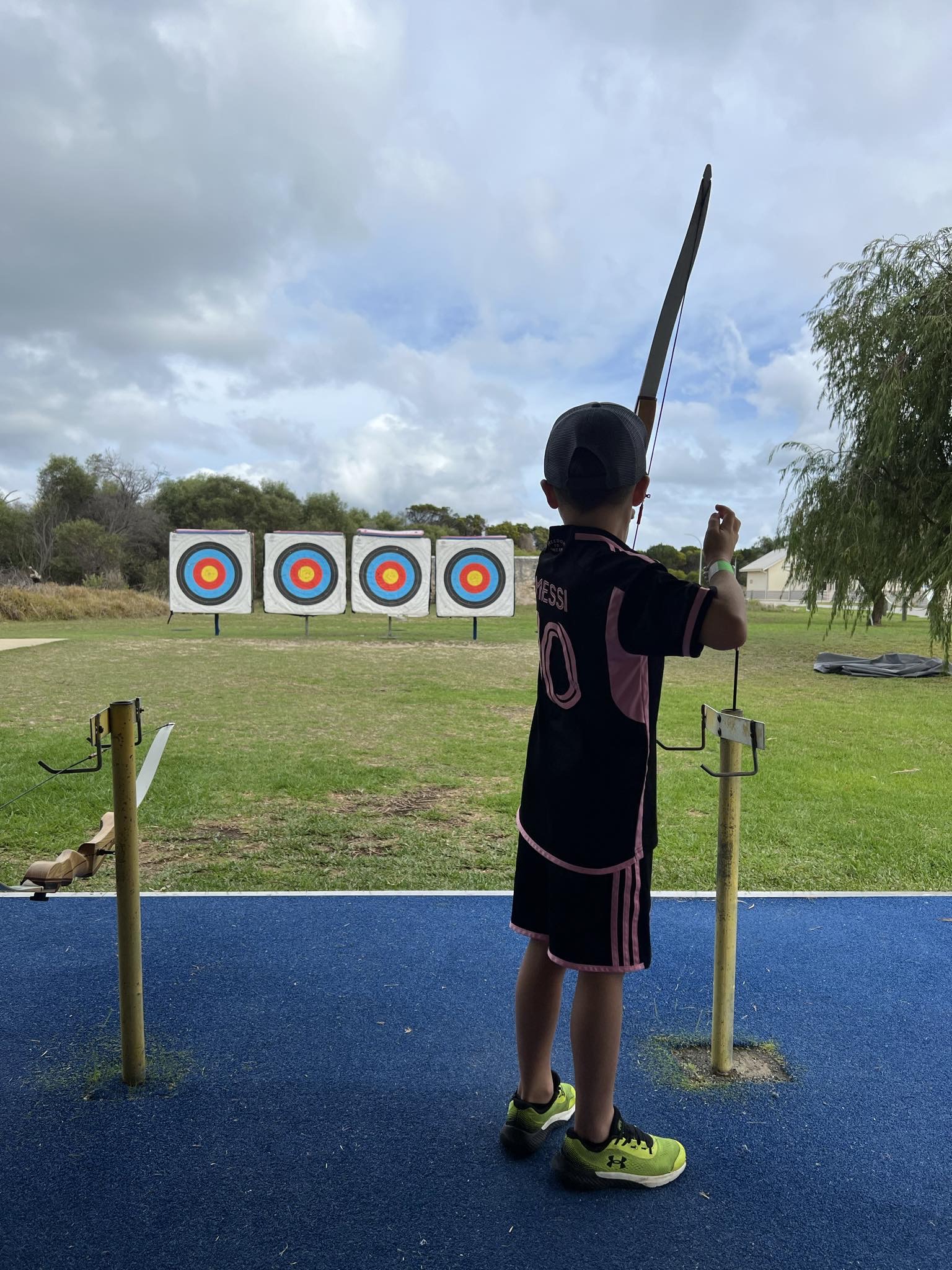 A boy in a black and red sports jersey stands at an outdoor archery range, holding a bow and aiming at round targets in the distance