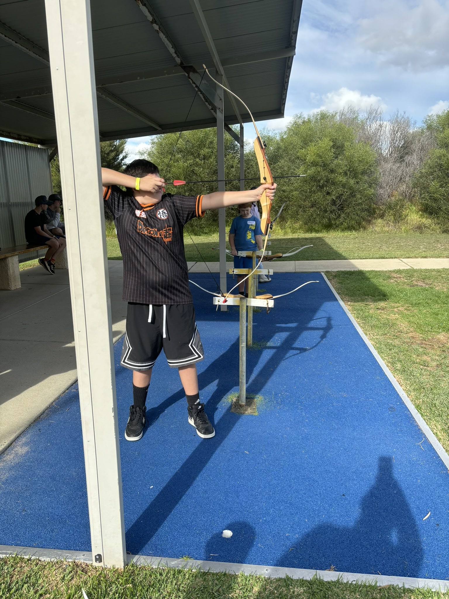 A boy wearing a black t-shirt and shorts, aiming his bow at a target