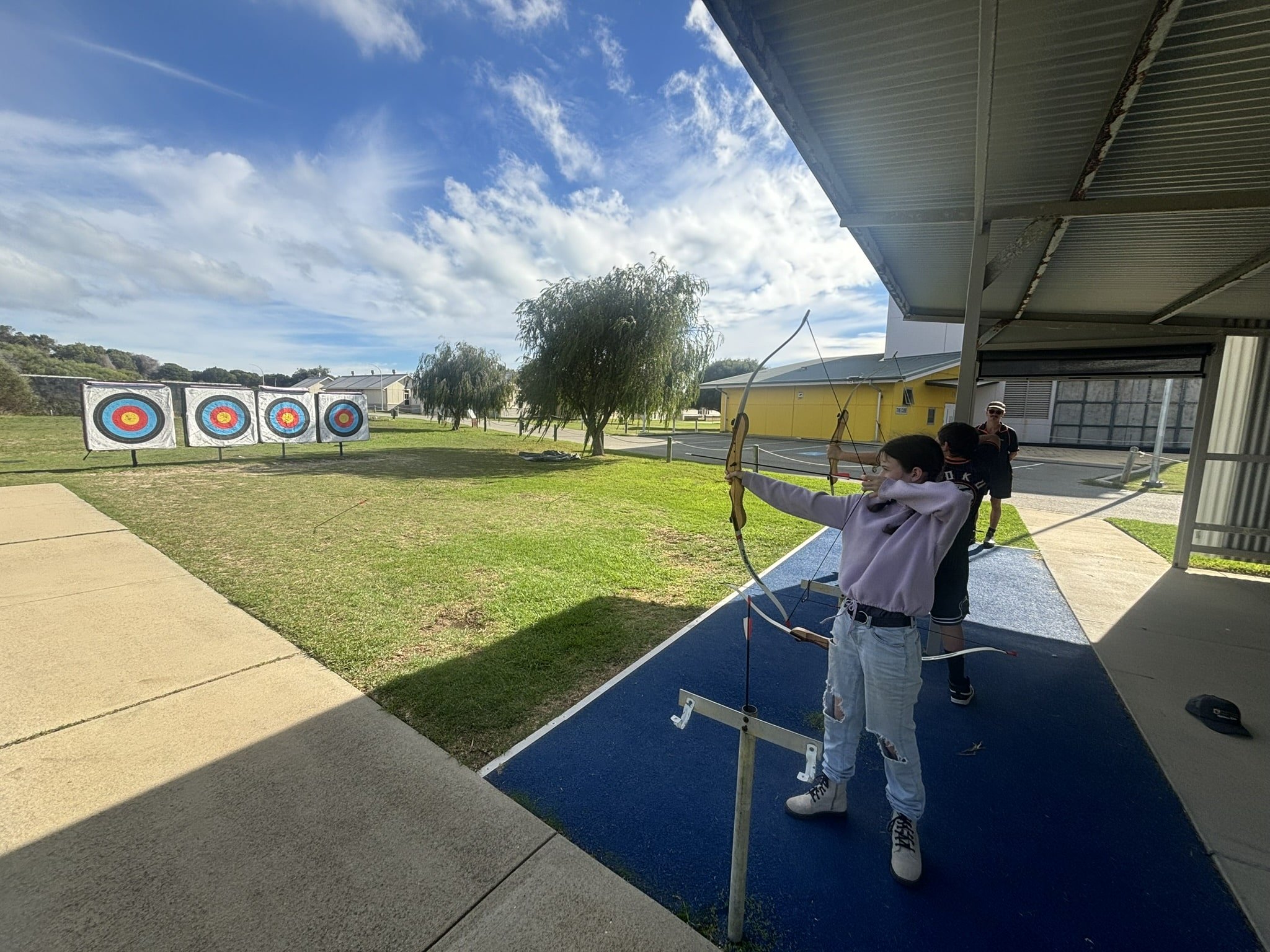 A girl in a lavender hoodie and jeans drawing back a bowstring as she prepares to shoot at an archery target