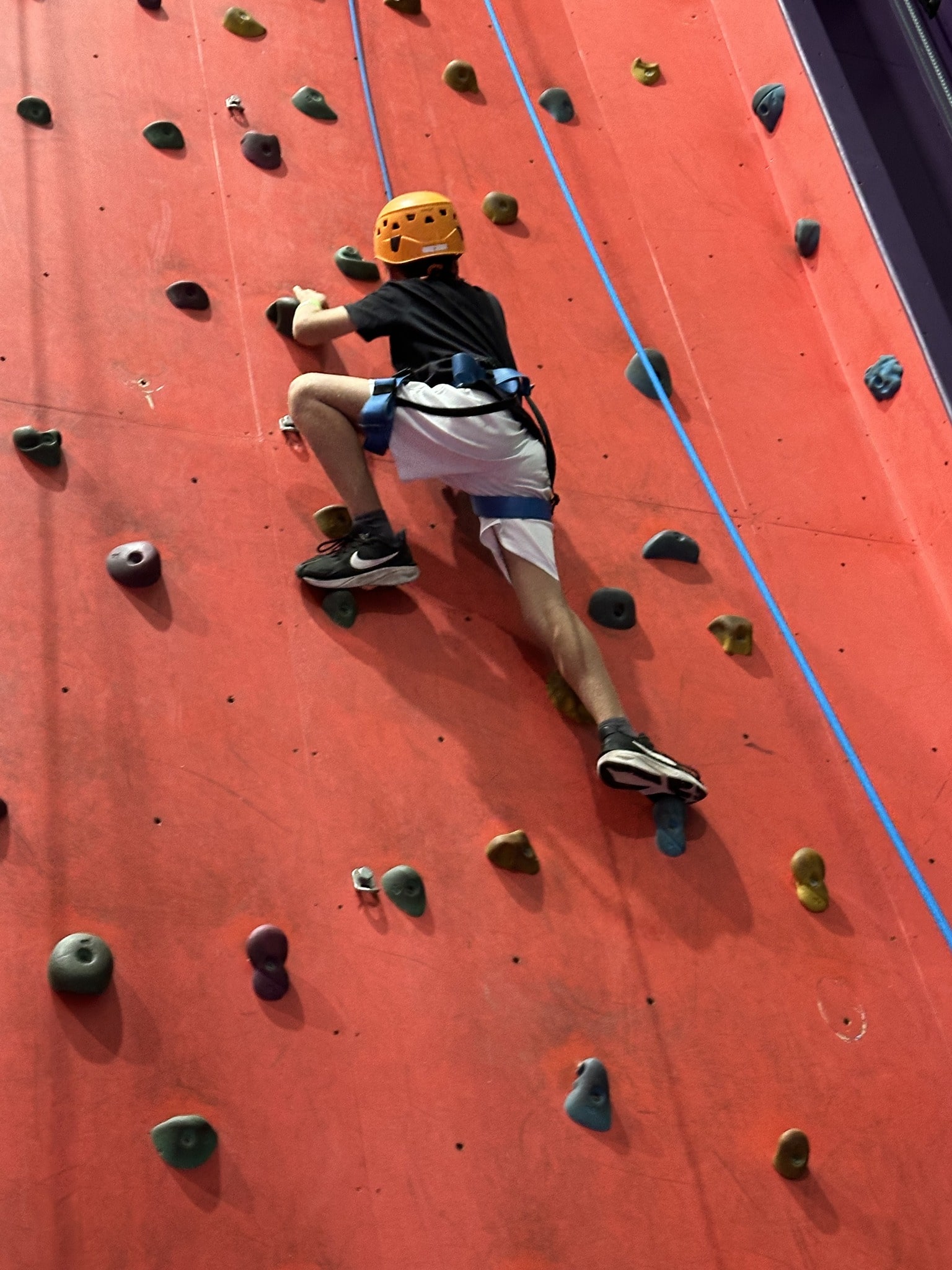 A boy wearing a helmet and harness climbs an indoor rock wall with red panels