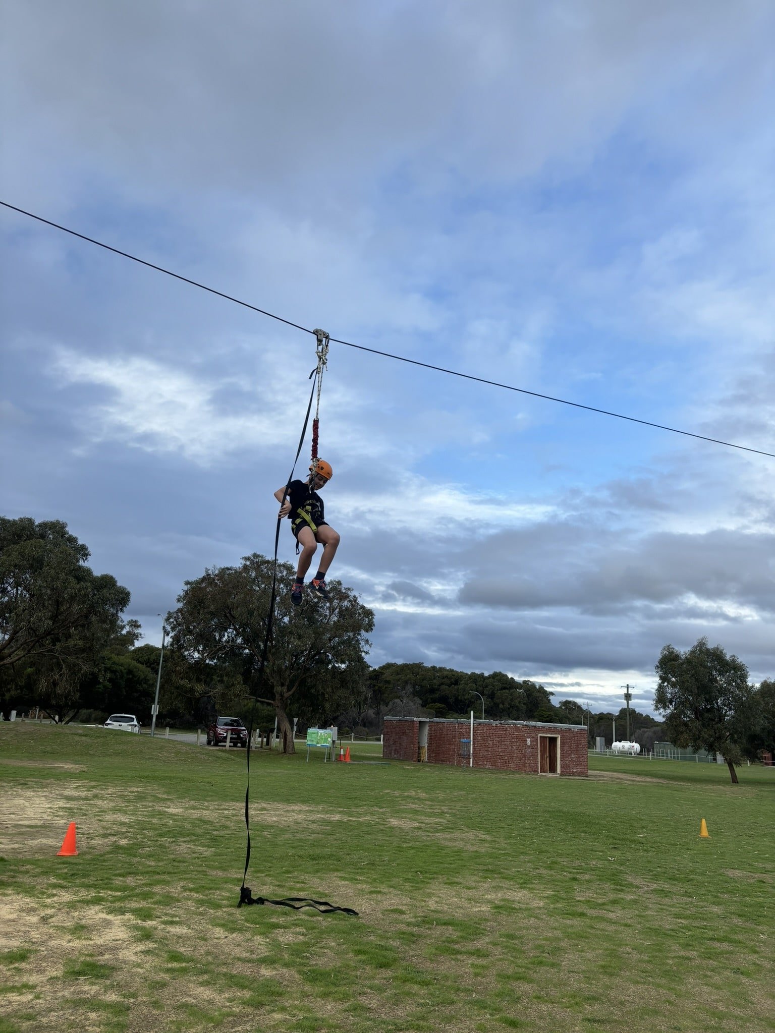 A child wearing a helmet and harness glides down a zip line over a grassy field