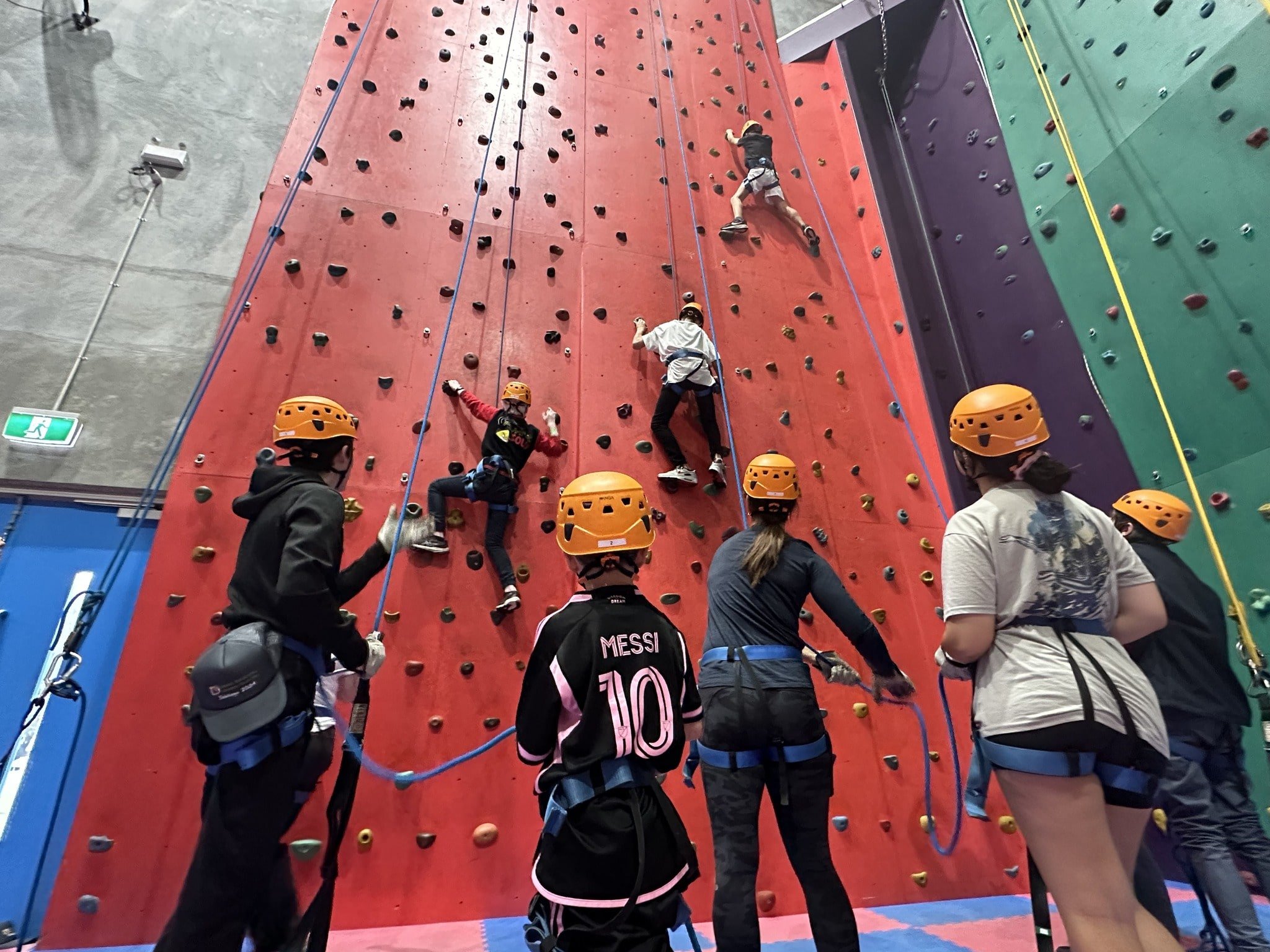 A group of children wearing helmets and harnesses stand in front of an indoor rock-climbing wall, watching others climb