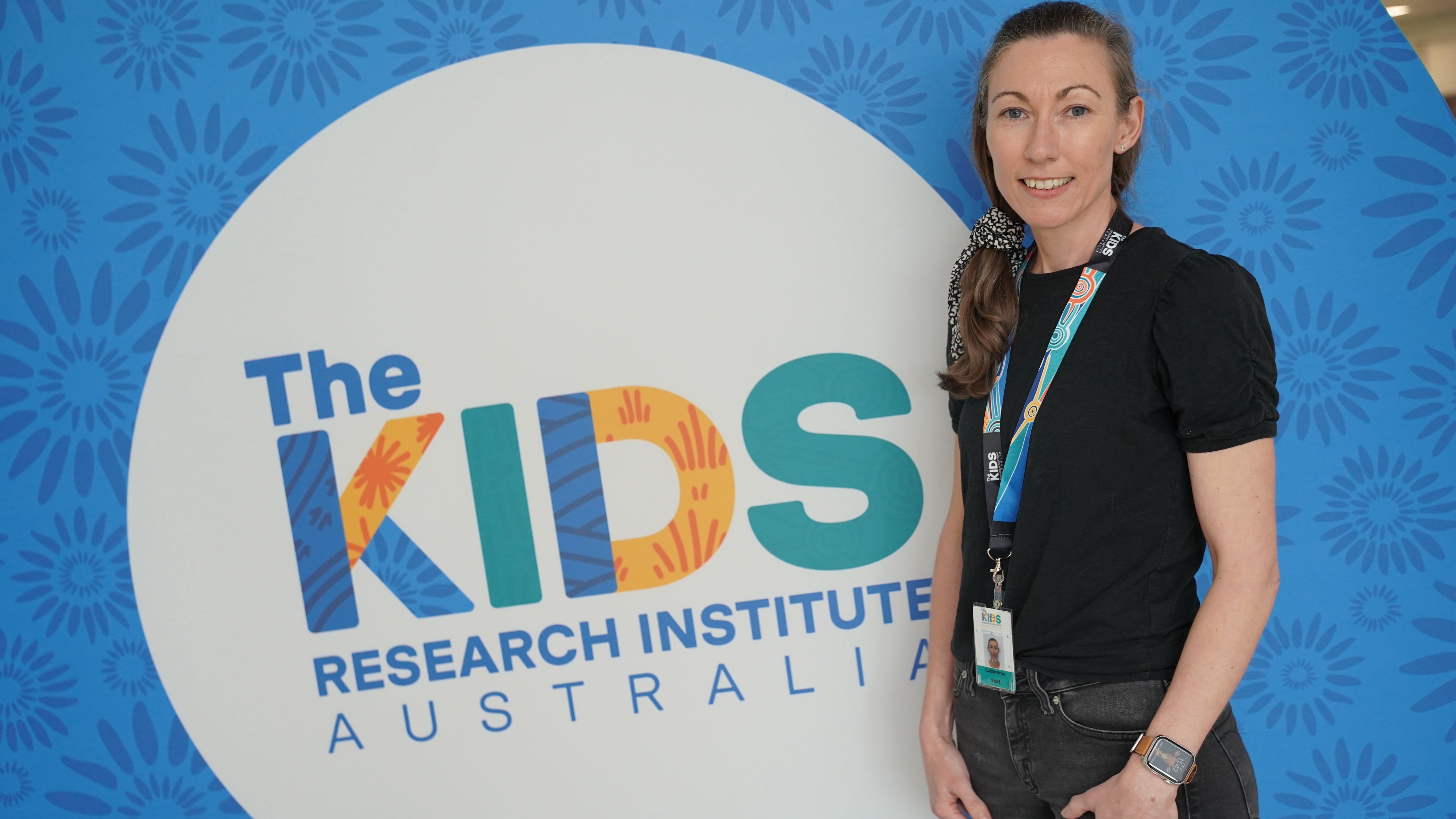 The Sibling Project PhD student, Caitlin Gray standing in front of The Kids logo