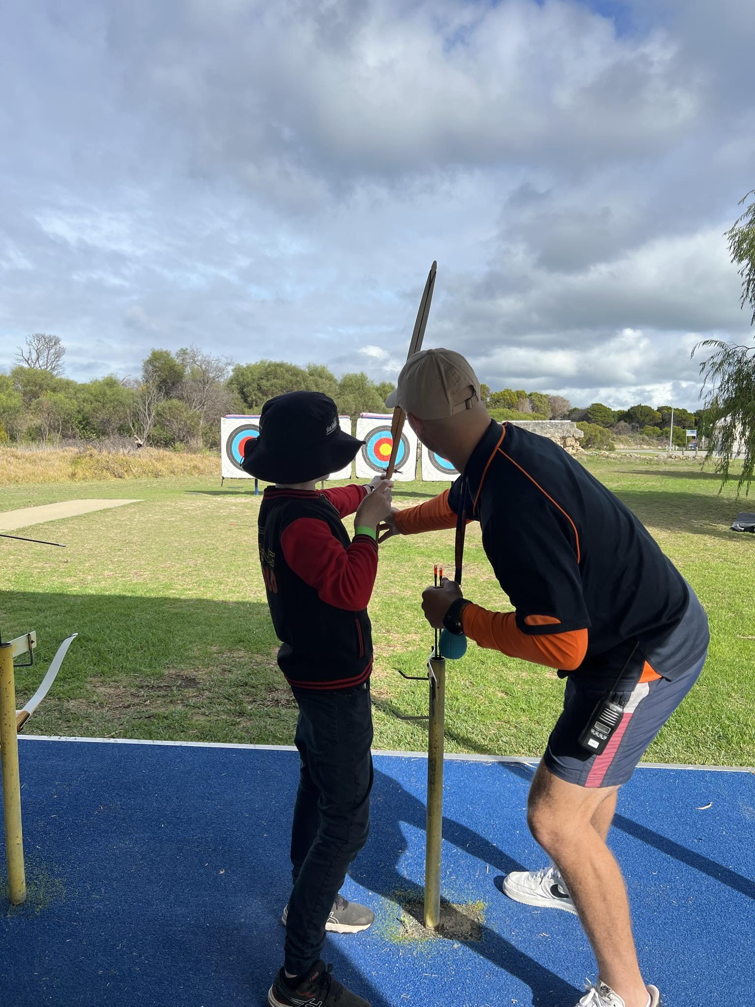 A young person wearing a black bucket hat and a red and black jacket is practicing archery with guidance from an instructor